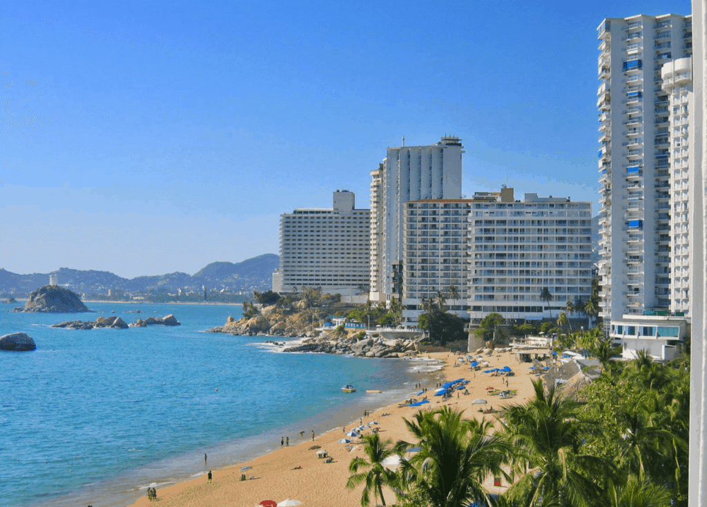 "Rocky cliffs overlooking Acapulco’s turquoise waters, with distant swimmers and sunlit waves breaking near the shore."