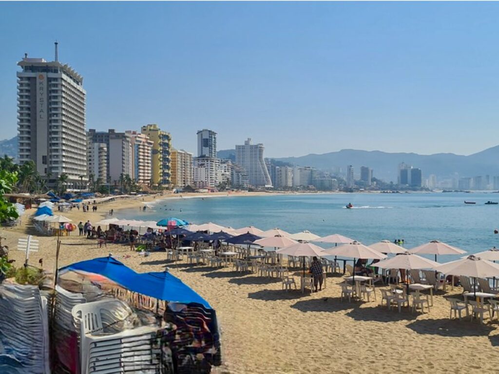 "Beachgoers relaxing under umbrellas on Acapulco’s main beach, with palm trees and vendors lining the sand."
